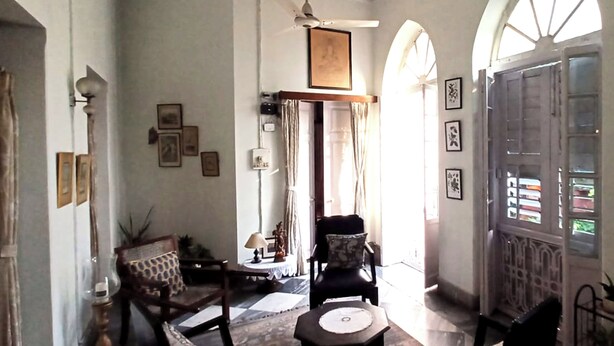 A spacious room with a high ceiling, a ceiling fan, and a large arched window. There are several framed pictures on the walls, a wooden chair, and a small table with a lamp. - Urban Trippers Lal Bari Heritage Arindam (Kolkata)