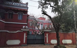 A red building with a gated entrance and a tree in front. - Urban Trippers Lal Bari Heritage Arindam (Kolkata)
