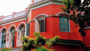 A red building with white trim and blue shutters, surrounded by greenery. - Urban Trippers Lal Bari Heritage Arindam (Kolkata)