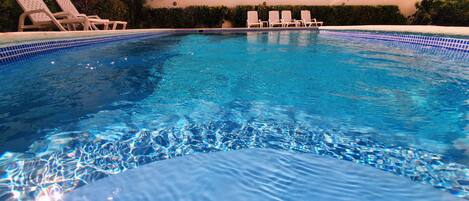 A swimming pool with clear blue water, surrounded by lounge chairs and a tiled edge.