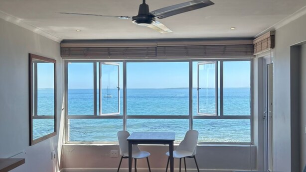 Ein Zimmer mit großem Fenster und Meerblick, einem Deckenventilator und einem Tisch mit zwei Stühlen.
