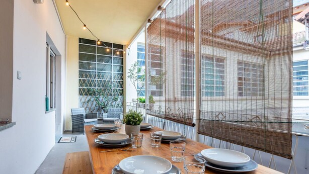 Un'area pranzo con un lungo tavolo in legno apparecchiato per quattro persone, circondata da ampie finestre che si affacciano su edifici e una pianta.