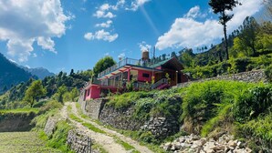 A rustic mountain house with a red exterior, a stone pathway leading to it, and a clear blue sky with scattered clouds.