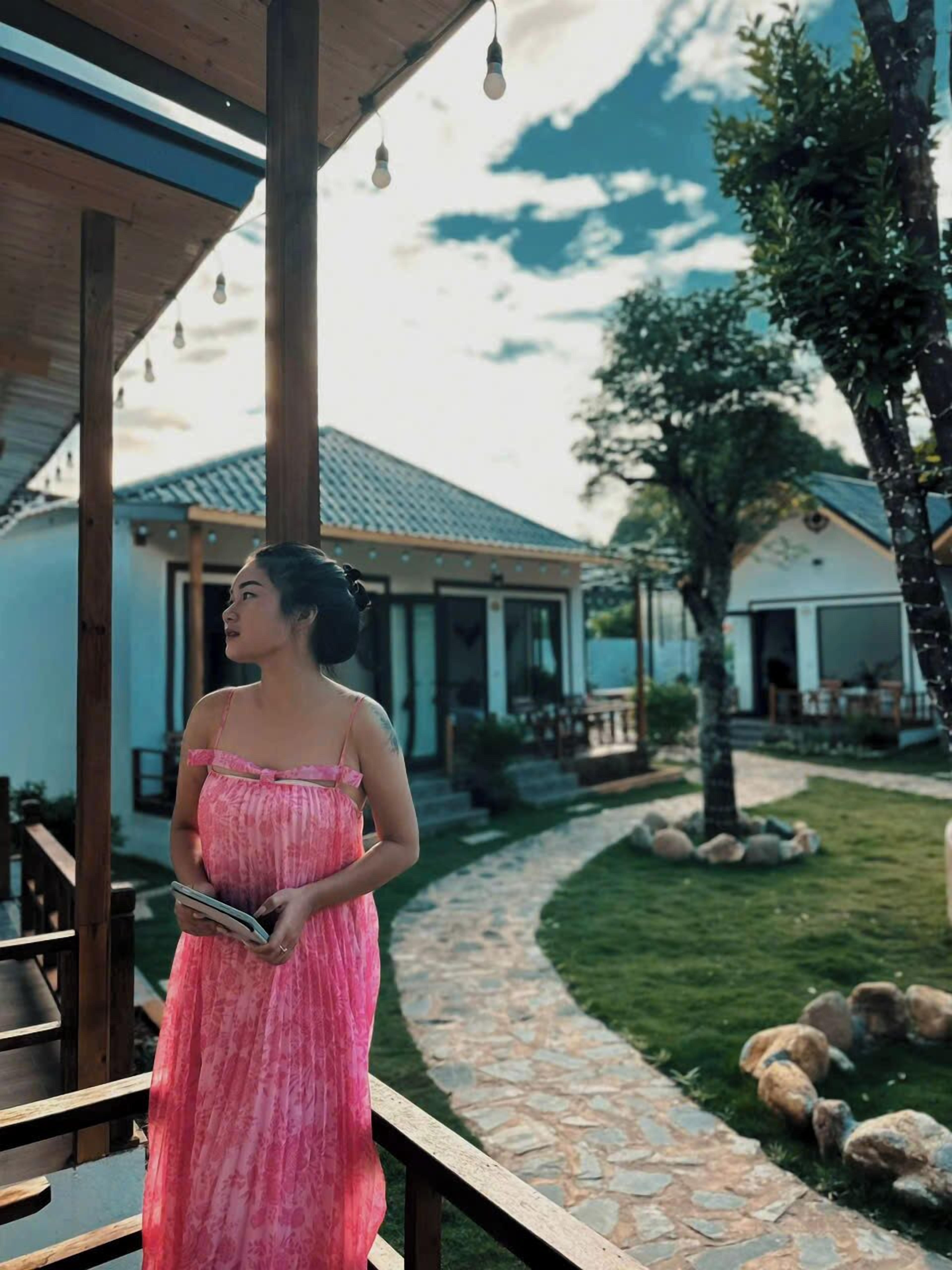 A woman in a pink dress standing on a wooden deck with a stone pathway leading to a building.