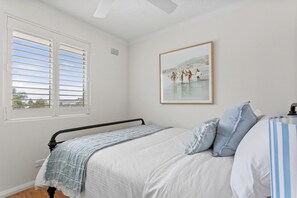 Ein Schlafzimmer mit einem Bett, einem Fenster mit Jalousien, einem gerahmten Bild an der Wand und einem gestreiften Kopfkissen.