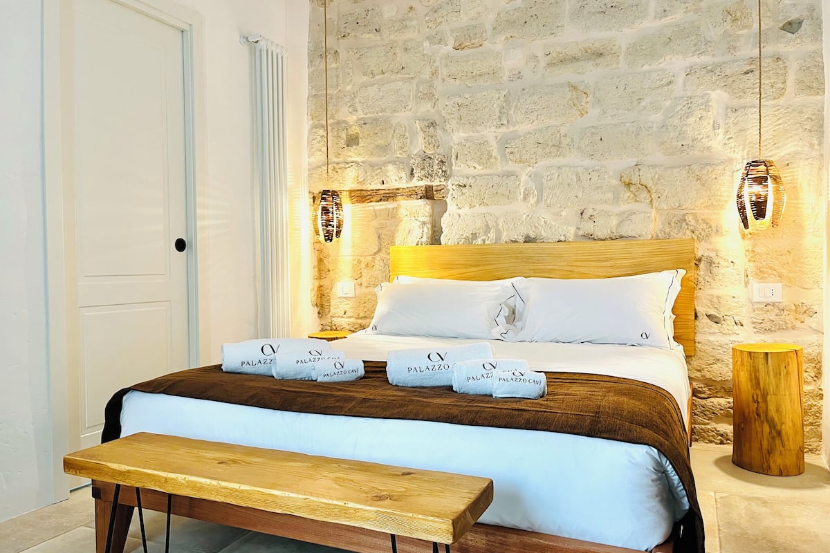 Une chambre avec un mur en pierre, un lit en bois, un banc et une table de chevet.