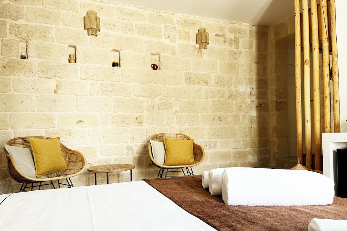 Une chambre d’hôtel avec un mur en pierre, deux fauteuils en osier avec des coussins jaunes, un lit avec du linge de lit blanc et un plaid marron.