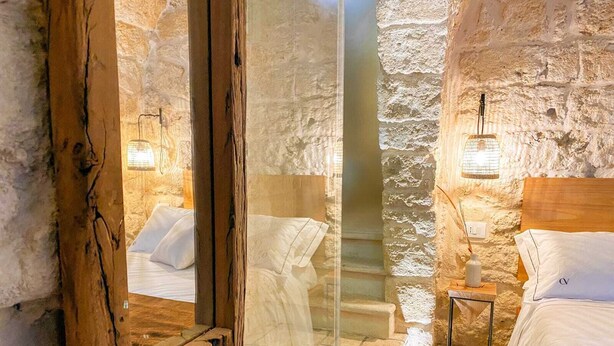 A stone-walled room with a wooden door, a bed with white bedding, a nightstand with a lamp, and a small table with a vase. - Palazzo CAVÌ  (Mola di Bari)