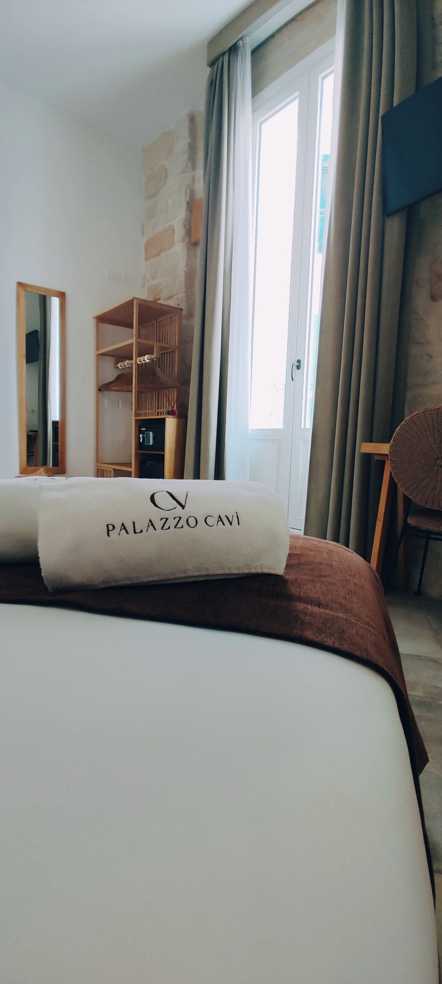 Una habitación de hotel con una cama bien hecha, un armario de madera, un espejo y vistas al exterior a través de una ventana con cortinas.