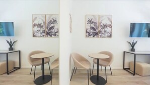 Een moderne kamer met twee ronde tafels, beige stoelen en zwarte metalen frames. Er zijn twee aan de muur bevestigde televisies en twee ingelijste botanische prints.