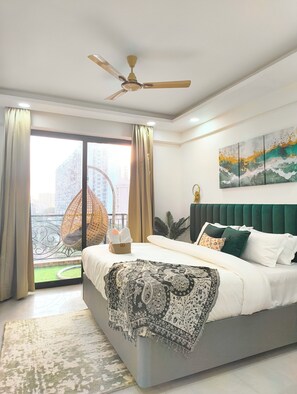 A modern bedroom with a large bed, a ceiling fan, and a view of a cityscape through the window. - Arth Corporate Suites at Gomti Nagar Ext (Lucknow)