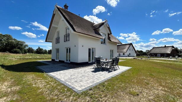 Kaksikerroksinen talo, jossa on olkikatto, kivetty patio ulkokalusteineen ja nurmikkoalue.