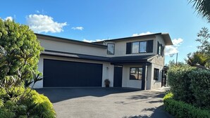 A modern two-story house with a garage, surrounded by lush greenery and a clear blue sky. - Coastal family home, 5 minutes walk to beach, hot tub, fully accessible (Papamoa)