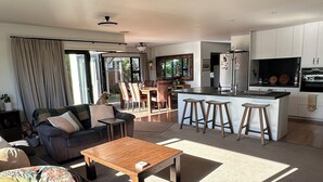 A spacious living room with a sofa, coffee table, and a dining area with chairs and a table. A kitchen with a large island and bar stools is visible in the background. - Coastal family home, 5 minutes walk to beach, hot tub, fully accessible (Papamoa)