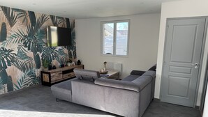 Un salon moderne avec un canapé d’angle gris, un téléviseur à écran plat fixé au mur, un papier peint à motifs tropicaux, une petite table d’appoint en bois et une porte grise.