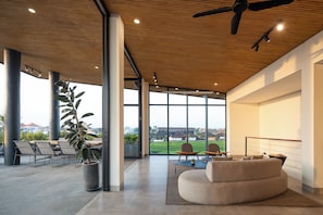 Un espacio interior amplio con ventilador de techo, ventanas grandes y una zona de estar con sofá y sillones.
