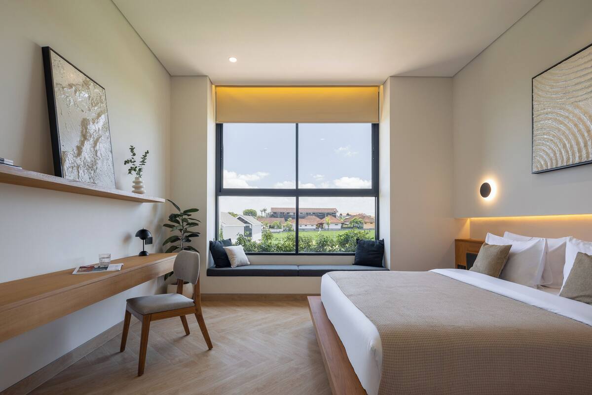 A modern hotel room with a large window, a desk with a chair, a bed, and a nightstand.