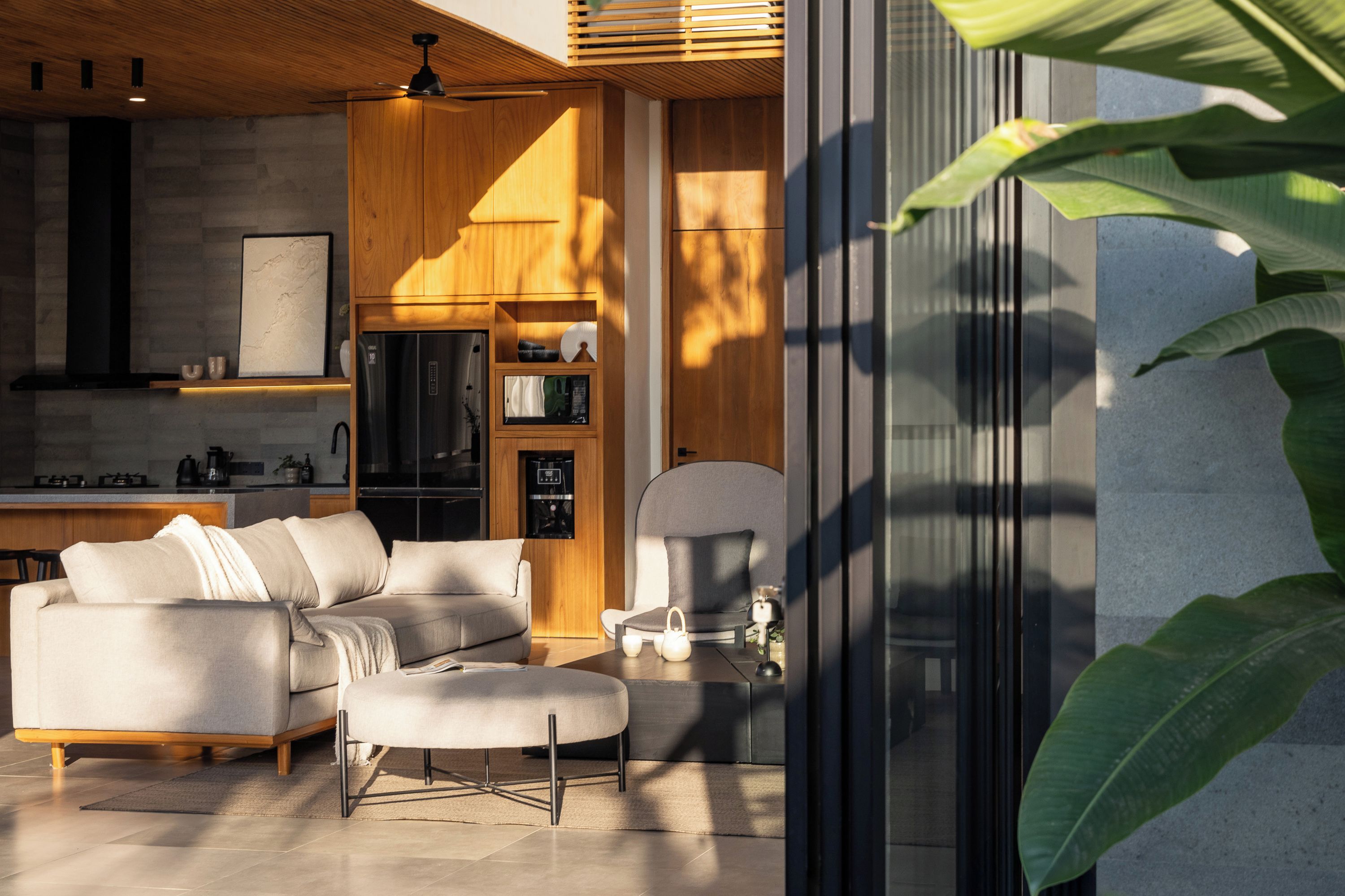 A modern living room with a sofa, ottoman, and a coffee table. There is a kitchen area with wooden cabinets and a refrigerator.