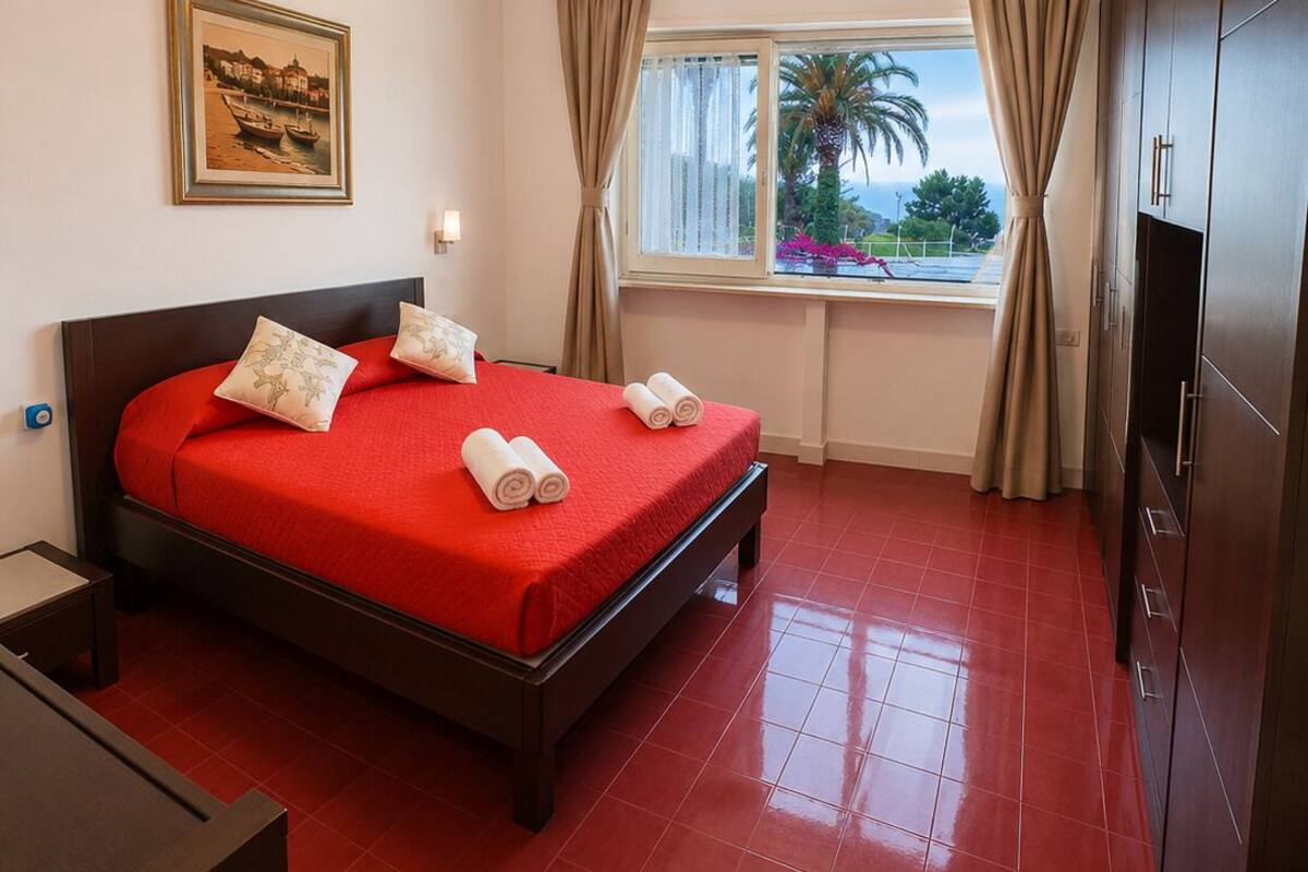 Una camera d'albergo con un letto, una scrivania, una finestra con tende, un quadro appeso al muro e una lampada a soffitto.