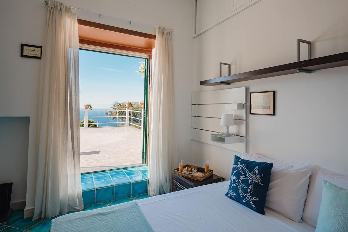 Camera d'albergo con un letto, un comodino, una mensola a muro e vista sul mare da un balcone.
