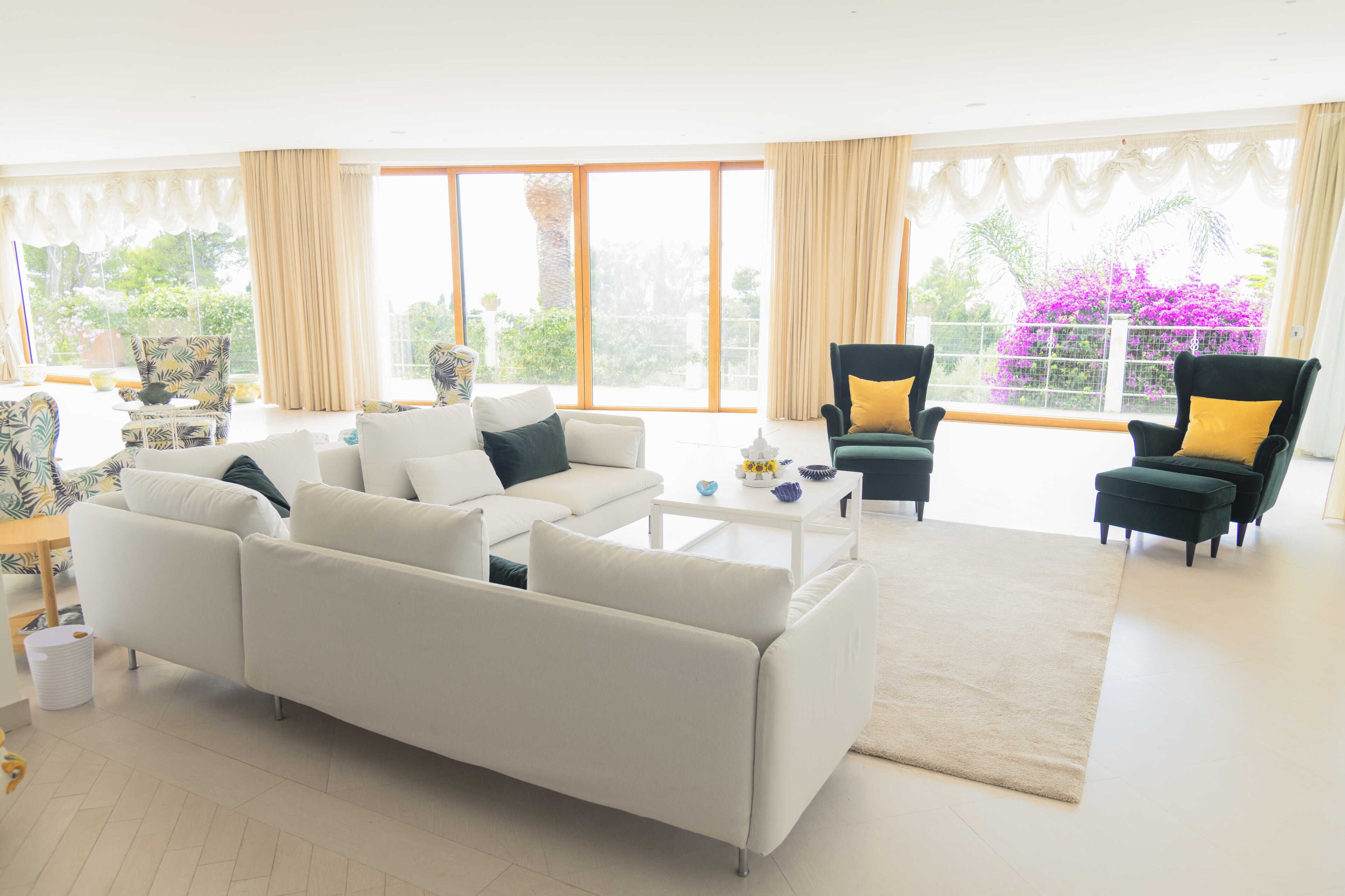 Un salón amplio con un sofá blanco, dos sillones y una mesa de centro. Ventanas grandes ofrecen vistas al exterior.