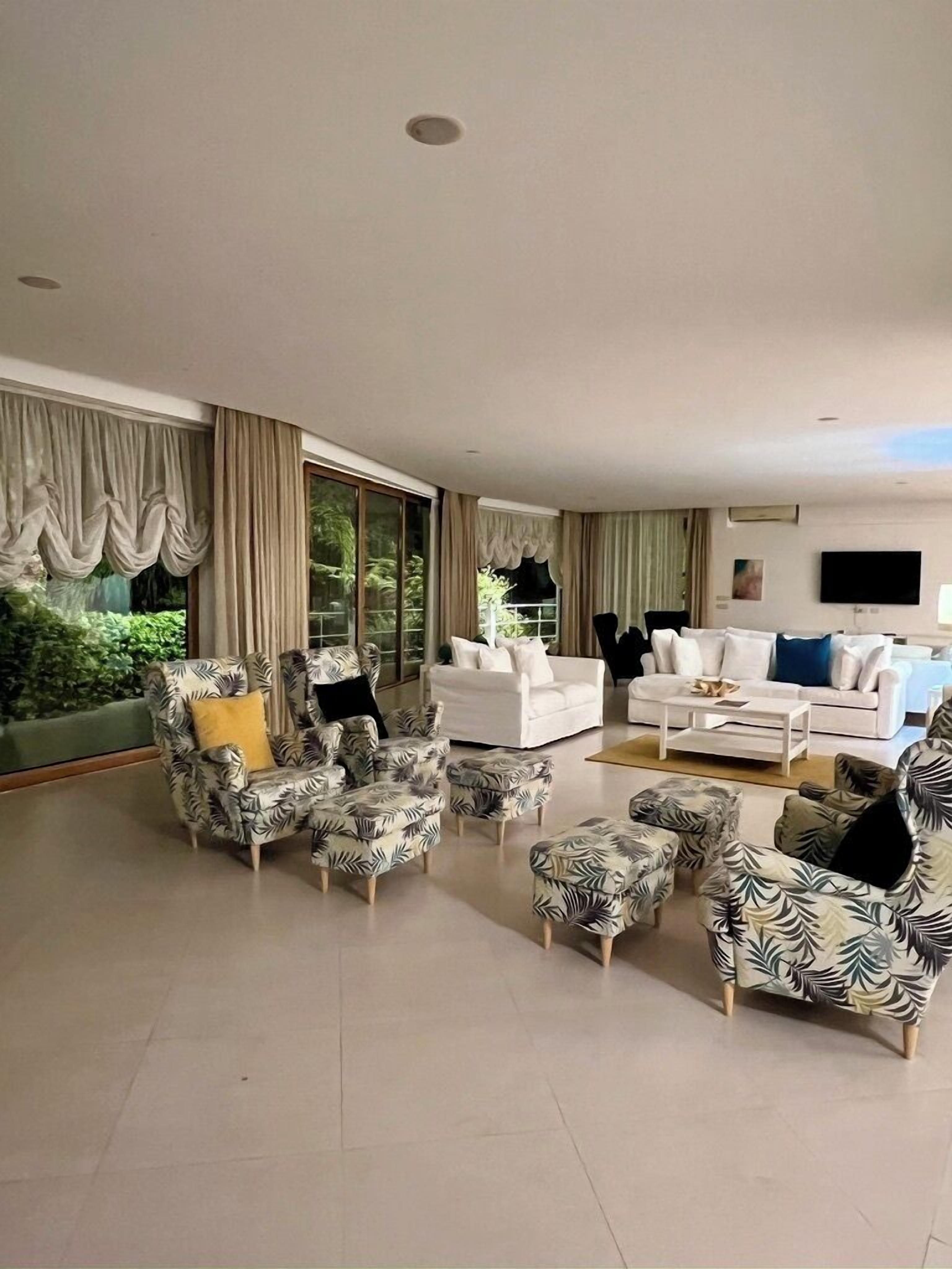 A spacious living room with a large window, a flat-screen TV mounted on the wall, and a variety of seating options including patterned armchairs and a white sofa.