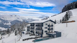 Un moderno edificio a più piani con balconi, piscina e montagne innevate sullo sfondo.