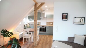 A modern kitchen with wooden beams, a dining area with white chairs, and a living room with a sofa and a plant. - Fewo zum Königreich.... where others go on vacation  (Westerwalsede)