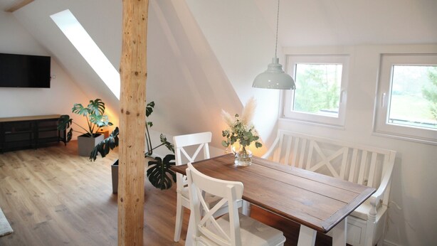 A dining area with a wooden table and white chairs, a hanging light fixture, and a view of the outdoors through windows. - Fewo zum Königreich.... where others go on vacation  (Westerwalsede)