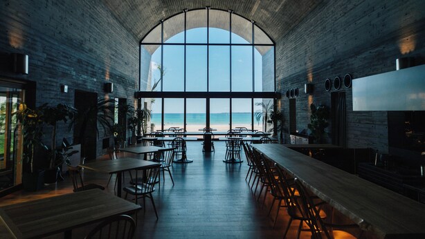 A spacious indoor area with a high arched window overlooking a beach, featuring wooden tables and chairs, and potted plants. - Colorit GOTO ISLANDS (Goto)