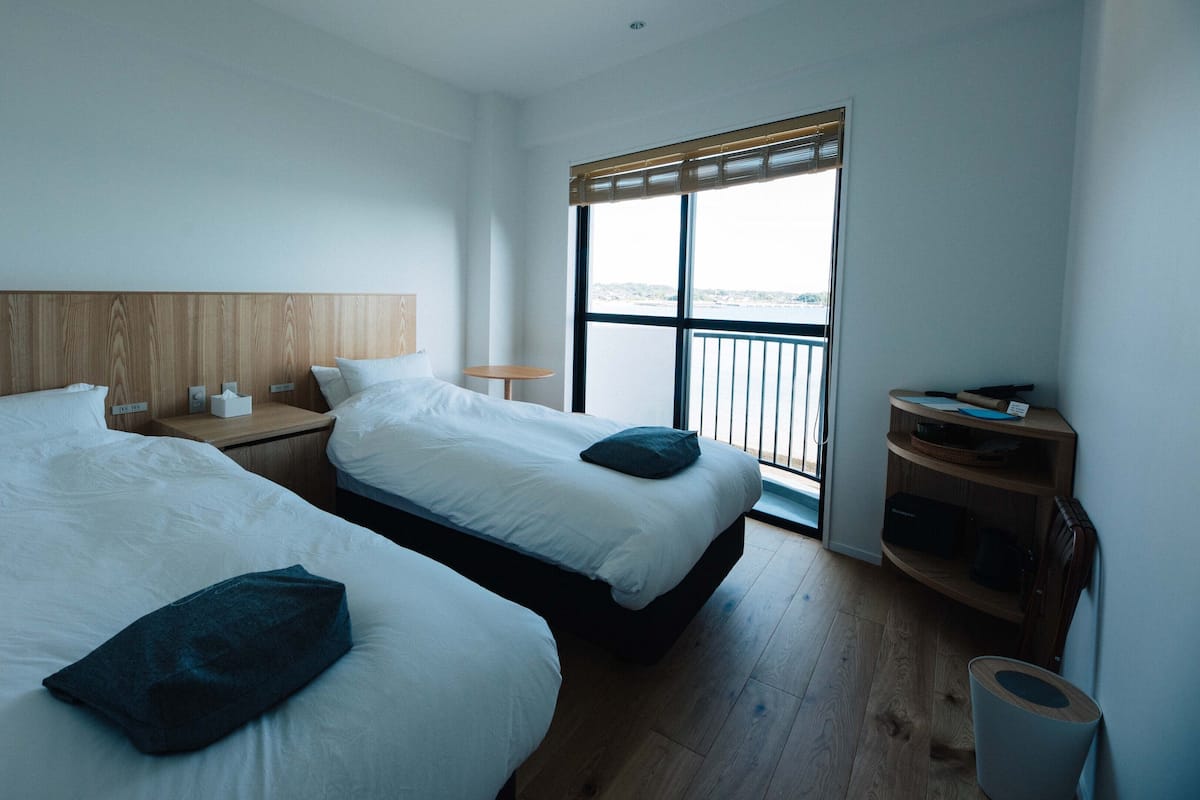 Habitación de hotel con dos camas, cabecero de madera, una mesita con una lámpara y vista al mar a través de un ventanal grande.