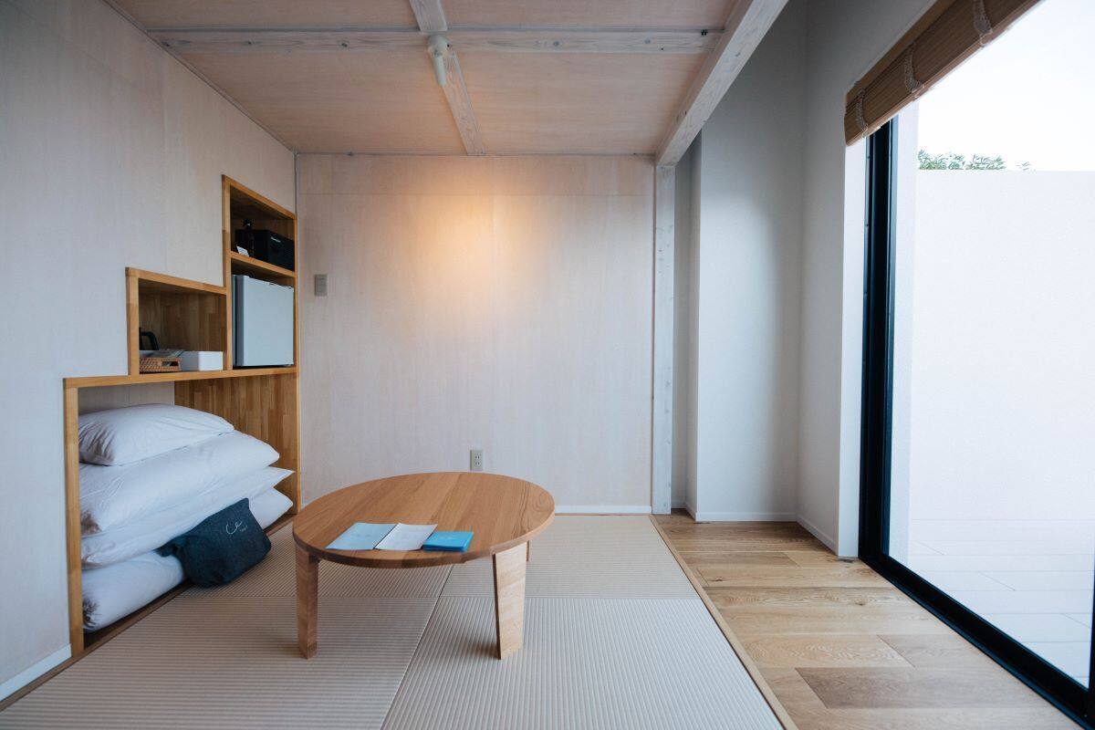 Una habitación minimalista con una mesa de madera, un estante empotrado con almohadas y un ventanal grande.