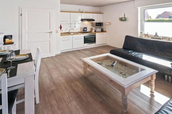 Uma sala de estar moderna com uma mesa de centro de vidro, um sofá de couro preto e uma área de jantar com uma mesa branca posta para dois.
