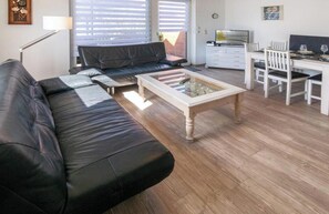 Una sala de estar con un sofá de cuero negro, una mesa de centro de madera, una zona de comedor con sillas blancas y un televisor sobre un mueble blanco.