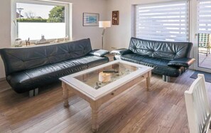Sala de estar com dois sofás de couro preto, uma mesa de centro de vidro, piso de madeira e vista pela janela.