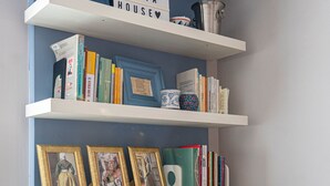 Um quarto com uma parede azul de destaque, uma estante branca com livros e quadros, uma TV de tela plana e uma placa que diz "WELCOME CUSTA HOUSE".