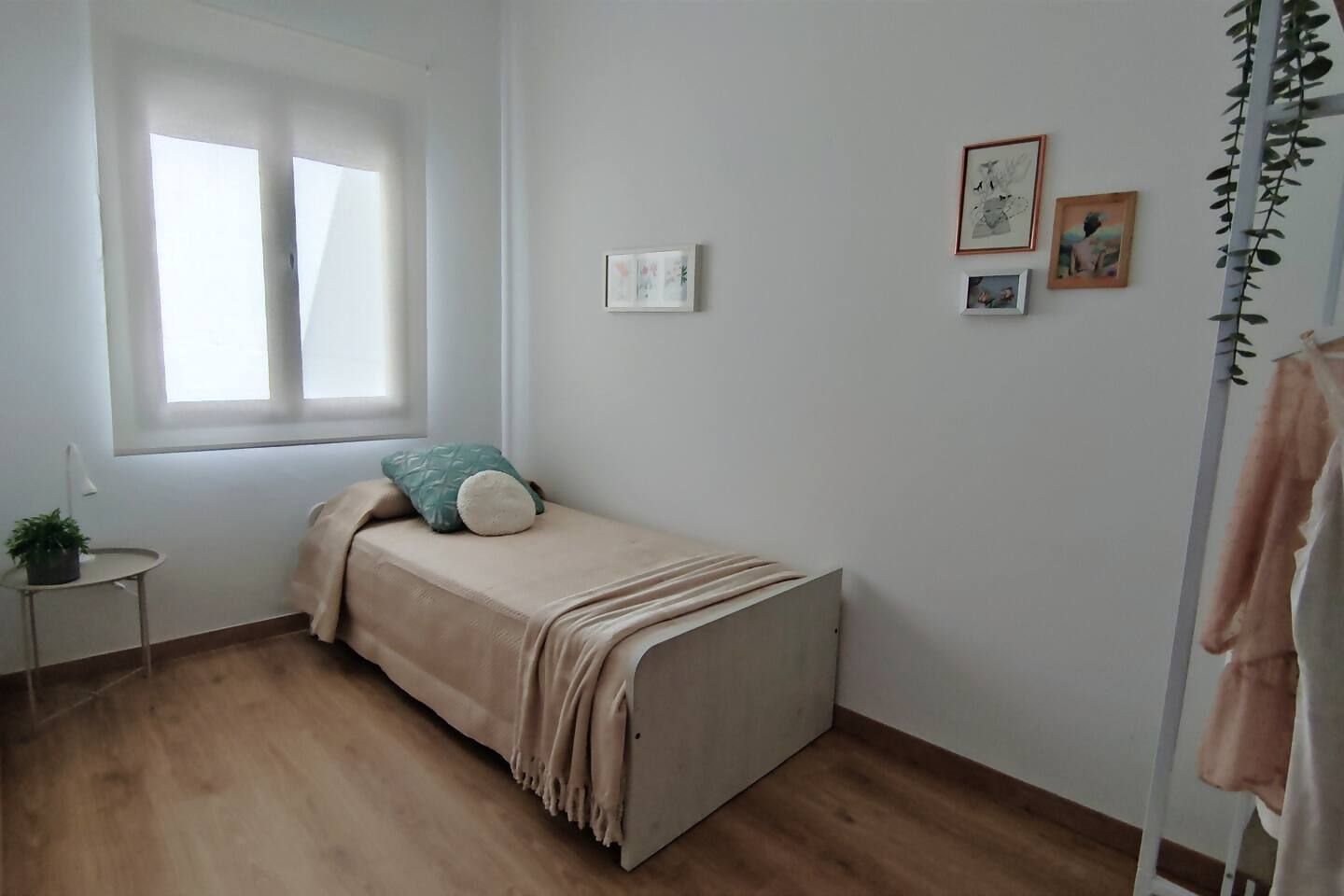Une petite chambre avec un lit simple, une fenêtre, une petite table avec une plante et quelques tableaux encadrés accrochés au mur.