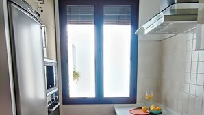 Cocina compacta con horno integrado, microondas y lavamanos. Se ve una ventana con persianas y una planta visible a través de ella.