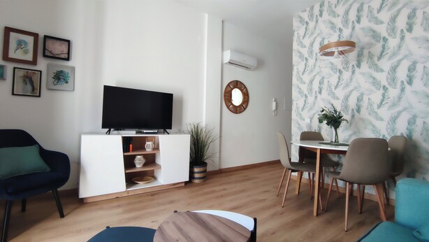 Una sala de estar con un televisor de pantalla plana sobre un mueble blanco, un comedor con una mesa redonda y sillas, y un papel tapiz estampado en una pared.