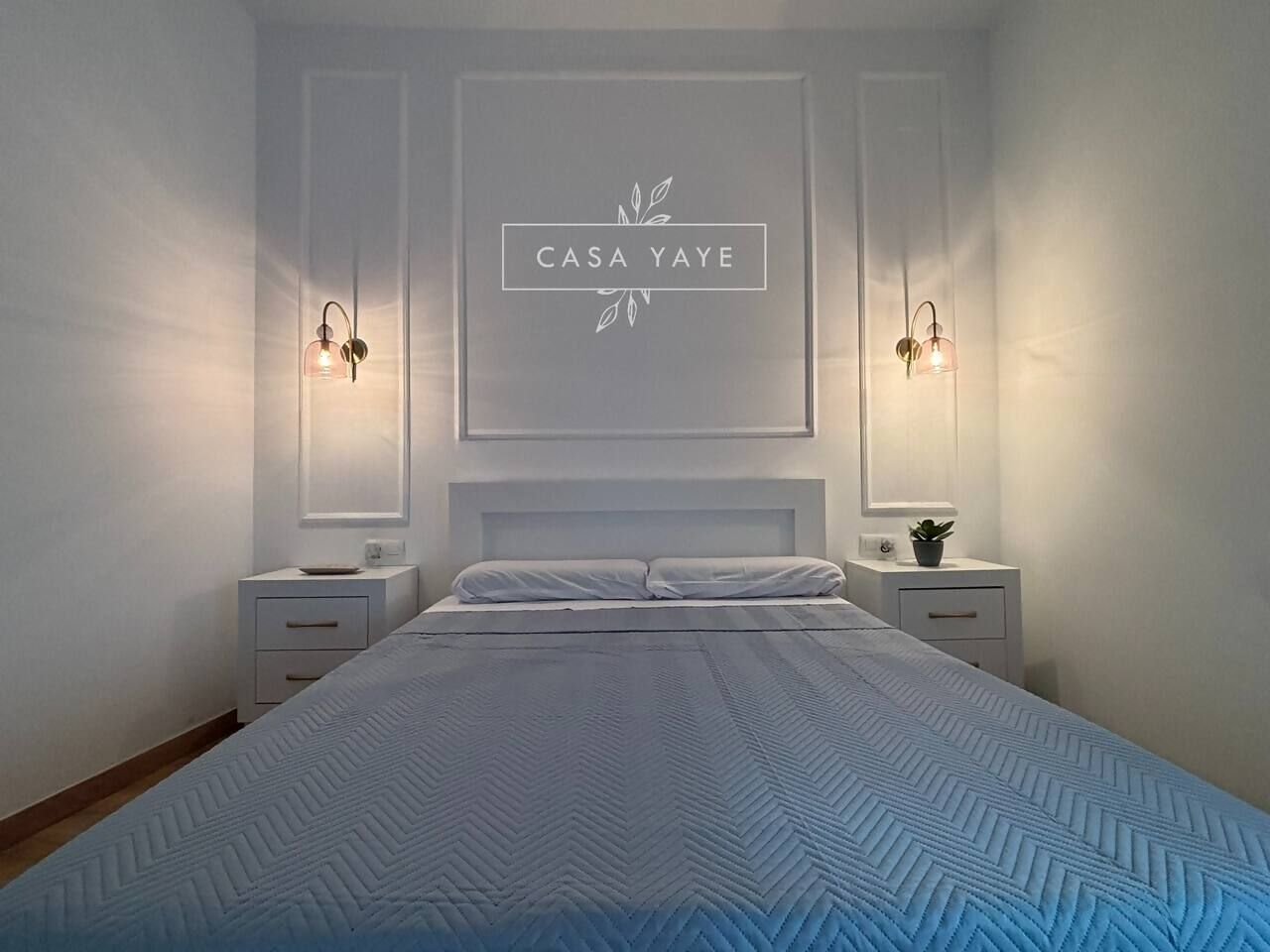 Un dormitorio con una cama, dos mesitas de noche, una lámpara de pared y un texto enmarcado en la pared.