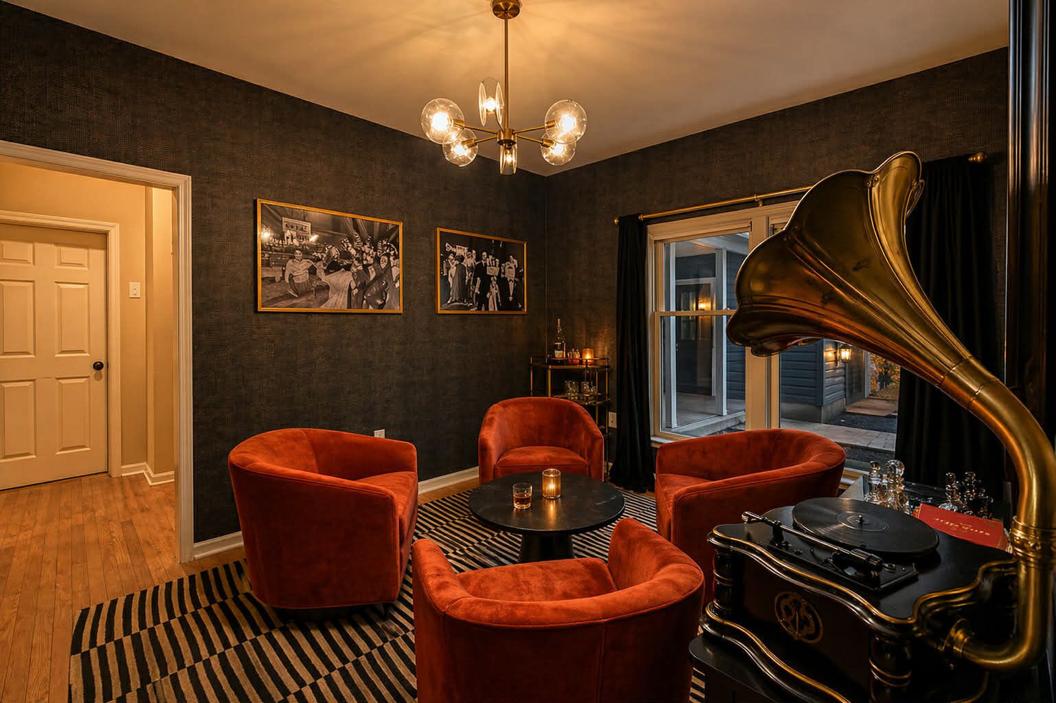A room with a black and white striped rug, a table with a glass, two red armchairs, a gramophone, and two framed black and white photographs on the wall.