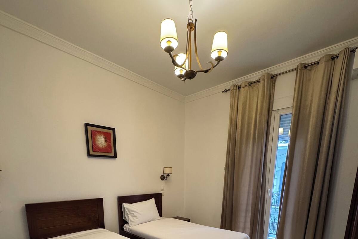 A hotel room with two single beds, a chandelier, a framed picture on the wall, and a view of the outside through the window.