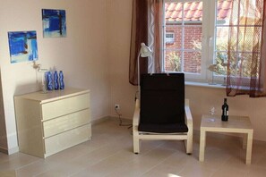 Relax in this serene space with a single chair by the window, a small table, and a dresser adorned with art.