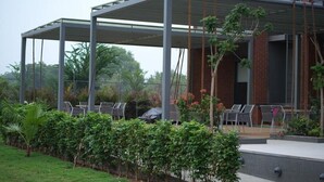 A covered outdoor seating area with chairs and tables, surrounded by greenery and a brick building. - FULLY FURNISHED STUDIO APARTMENT WITH   BALCONY,  (Vadodara)