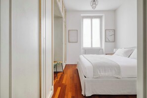 A bedroom with a wooden floor, a bed with white bedding, a window with white curtains, and a radiator.