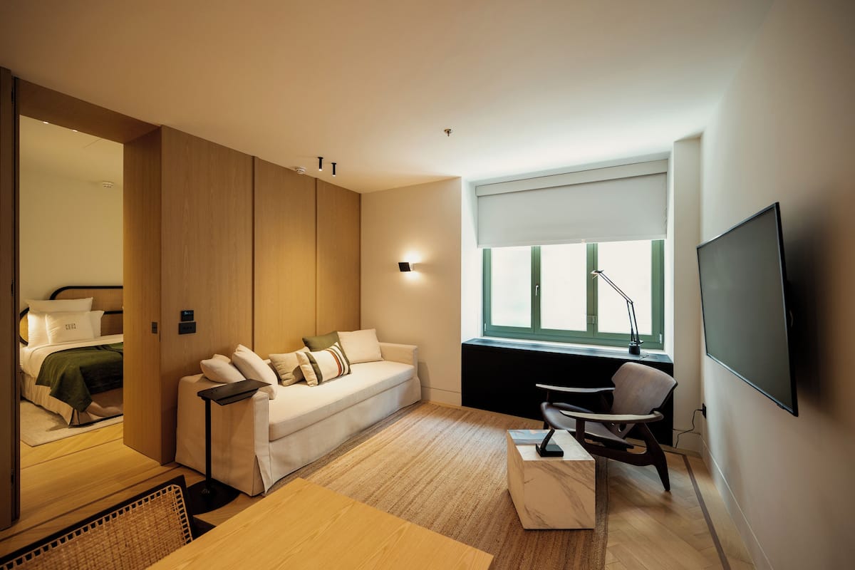 A modern hotel room with a sofa, a desk, a chair, a TV, and a window with blinds.