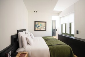 A hotel room with a bed, a desk with a laptop, a framed picture on the wall, and a window with blinds. - Athens Capital Suites-Mgallery Collection (Athens)