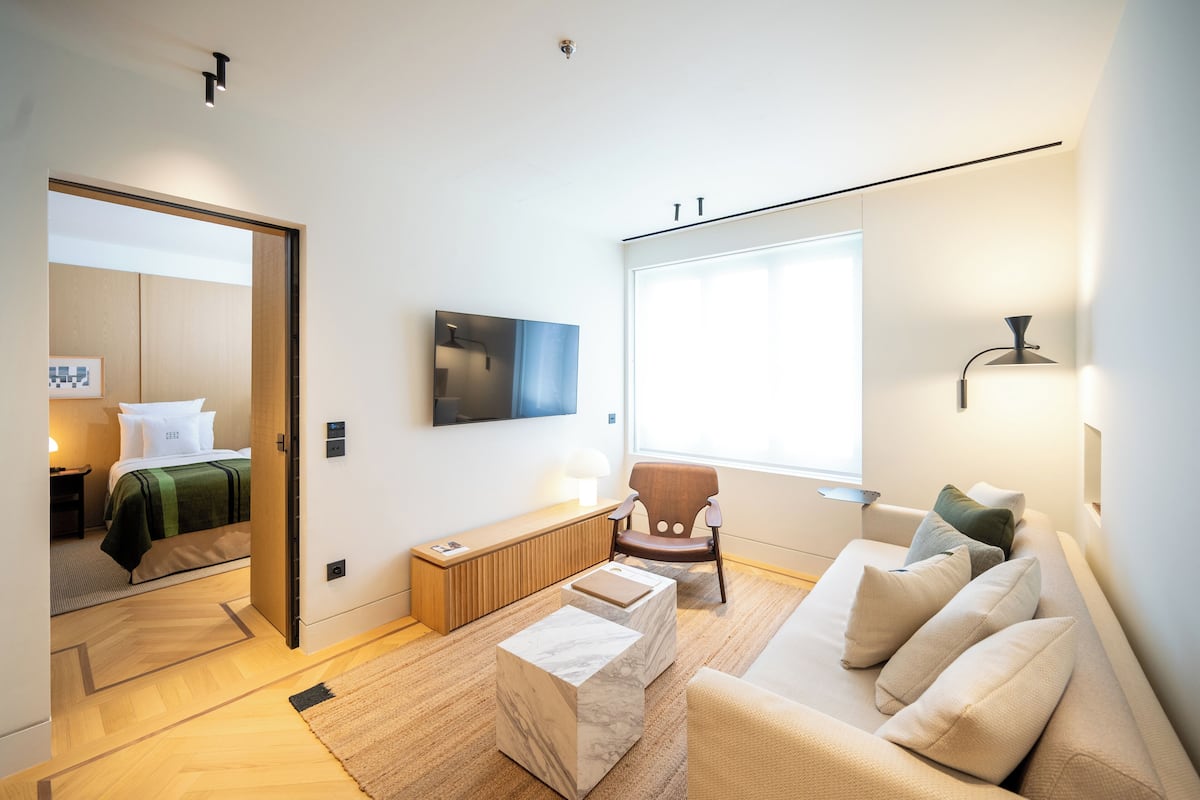 A modern hotel room with a bed, a sofa, a chair, a TV, and a window.