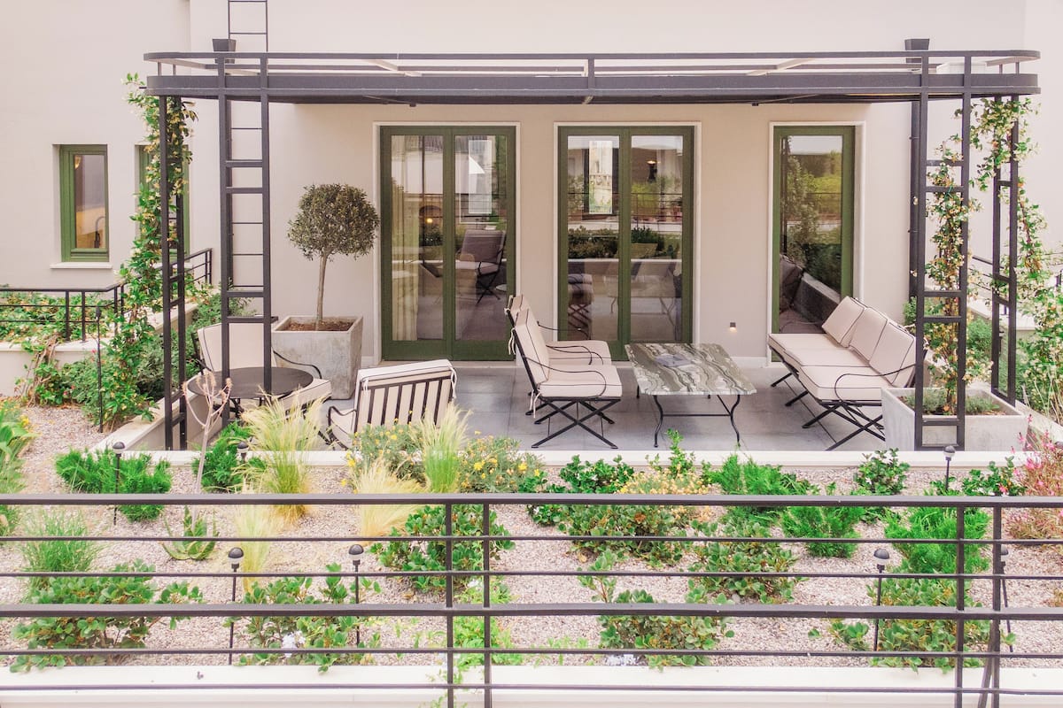 A modern outdoor patio with a dining table and chairs, surrounded by potted plants and a metal railing.