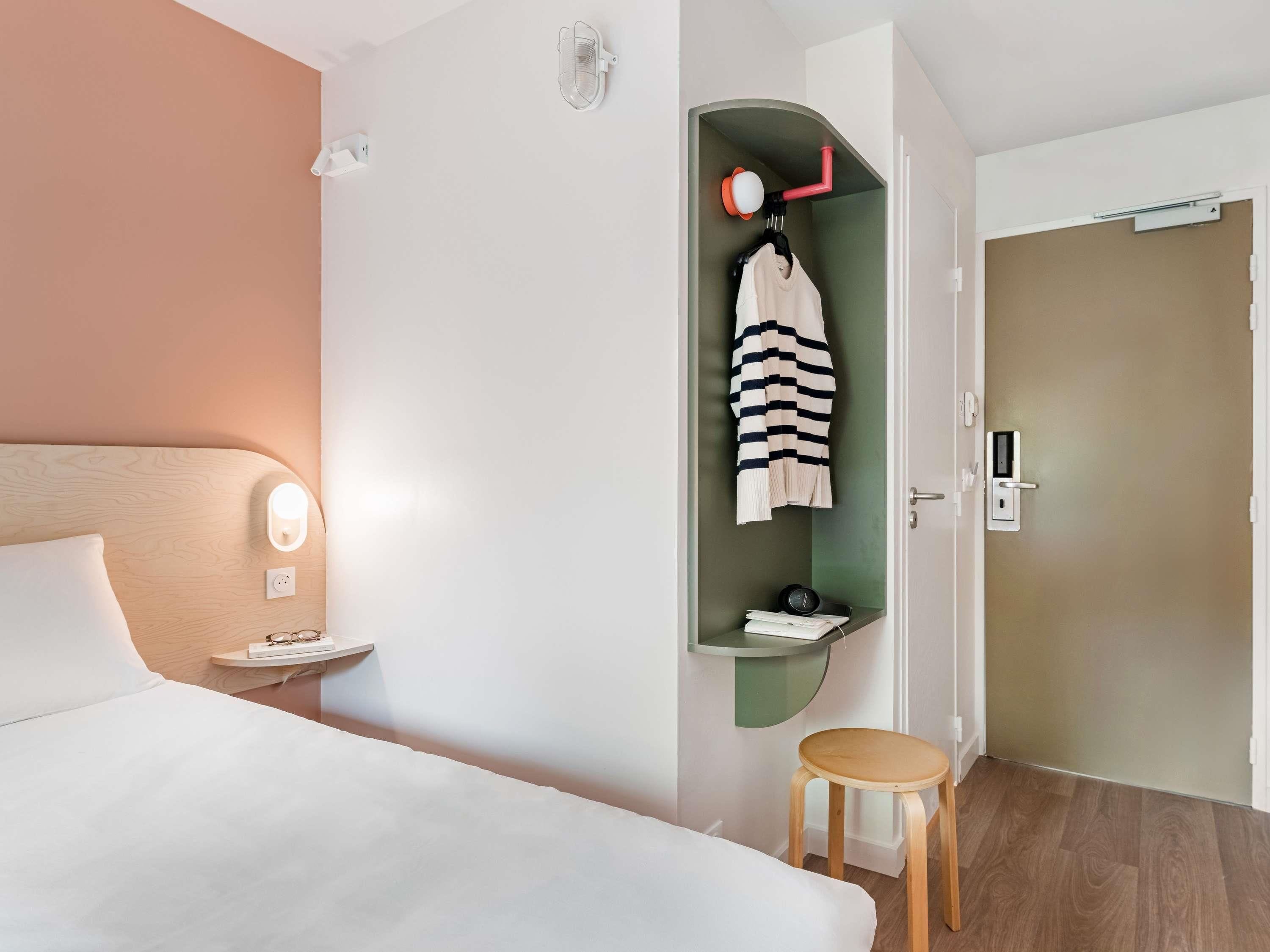Habitación de hotel con cama, mesita de noche, estante con lámpara de pared, repisa con una toalla y puerta con cerradura.